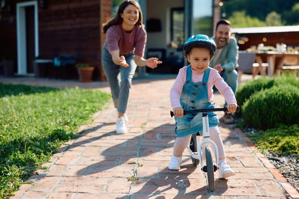 Small girl riding bicycle with the support of her parents outdoors.